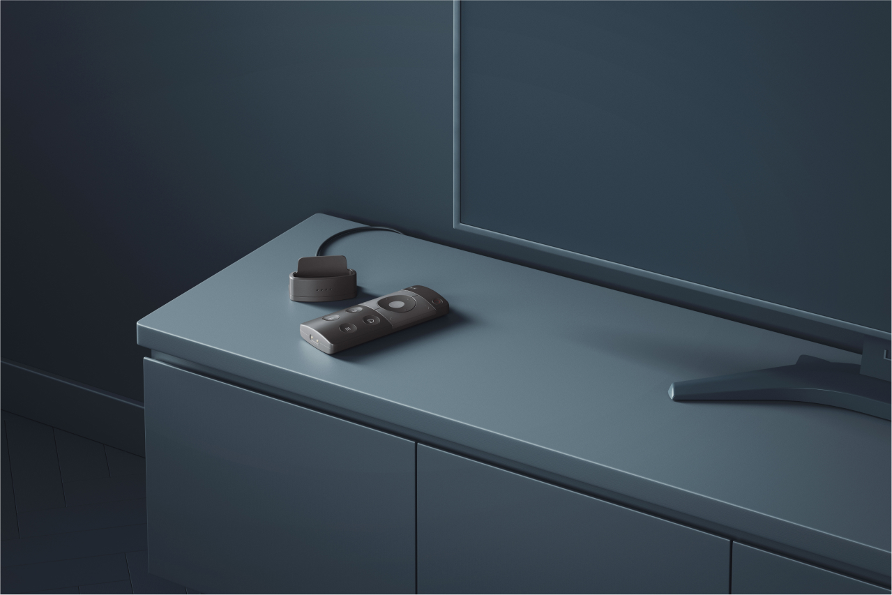 A sleek remote control placed next to its charging dock on a modern cabinet in a dark, minimalist setting.