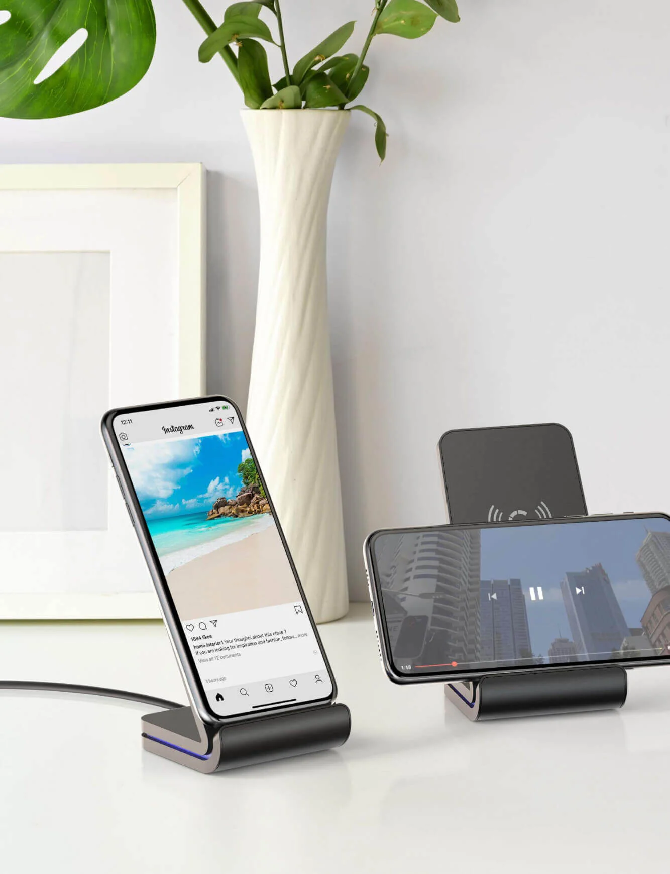 Two smartphones on wireless charging stands: one upright with a beach photo, the other sideways playing a video, placed on a white desk with a vase and plant.